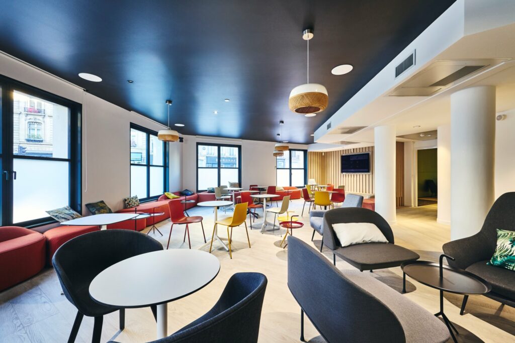 Agencement d'un espace détente modulable chez CCR, avec peinture foncée plafond, chaises et tables multicolores, banquettes en modules, alcôve sur mesure avec banquette et insert d'une télévision.