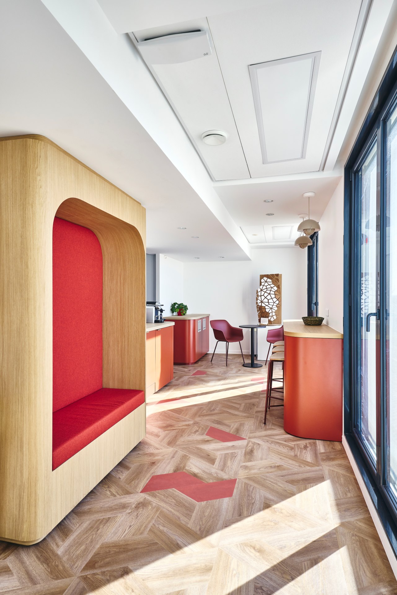 Agencement d'une kitchenette chez CCR, comprenant une alcôve menuisée sur mesure avec banquette intégrée, un bar sur mesure, un sol PVC effet bois posé en chevron déstructuré, des tabourets hauts et deux meubles plan de travail sur mesure.